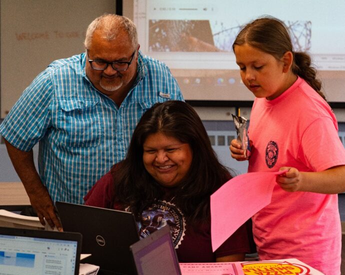 In this image, there are three people in what looks to be a classroom setting. On the left stands a man in a blue, button-up shirt with black glasses. In the middle, a woman sits in front of a computer in a maroon tshirt smiling at the screen. On the right side of the image is a young lady in a bright pink tshirt, holding a pink piece of paper. Credit: Indian Leader/Emmitt Brazille