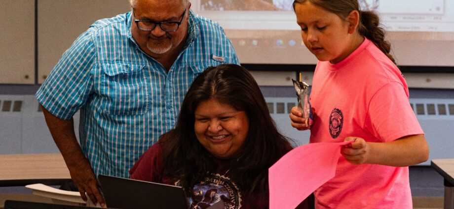 In this image, there are three people in what looks to be a classroom setting. On the left stands a man in a blue, button-up shirt with black glasses. In the middle, a woman sits in front of a computer in a maroon tshirt smiling at the screen. On the right side of the image is a young lady in a bright pink tshirt, holding a pink piece of paper. Credit: Indian Leader/Emmitt Brazille